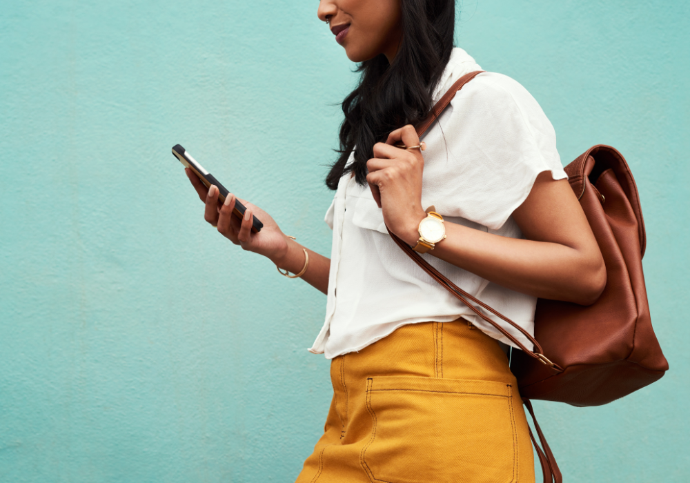 Young woman walking while looking at her phone, wearing a white blouse, mustard yellow trousers, and a brown backpack, agains