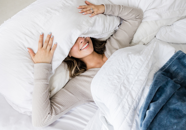 A woman smiling while lying in bed, partially covering her face with a white pillow.